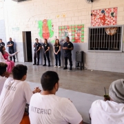 Grupo de detentos com camisetas brancas escrito "INTERNO" sentados em volta de uma mesa, participando de uma roda de conversa em um pátio. À frente deles, uma mulher fala ao lado de agentes da Polícia Penal. As paredes ao fundo possuem pinturas coloridas de personagens de desenhos animados.