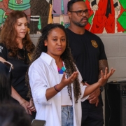 Uma mulher negra de cabelos cacheados e camisa branca fala de forma expressiva com as mãos abertas. Ao fundo, agentes da Polícia Penal e murais coloridos na parede do presídio.