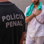 Em primeiro plano, as costas de um uniforme preto com a inscrição "POLÍCIA PENAL". Ao fundo, de forma levemente desfocada, um homem com vestes brancas e uma toalha verde no ombro está com a mão no coração.