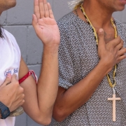 Detalhe aproximado de colares de contas (guias) em tons de verde, azul e branco sobre uma bata branca. Ao fundo, um homem usa um crucifixo de madeira, simbolizando a diversidade religiosa no local.