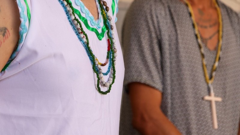 Detalhe aproximado de colares de contas (guias) em tons de verde, azul e branco sobre uma bata branca. Ao fundo, um homem usa um crucifixo de madeira, simbolizando a diversidade religiosa no local.